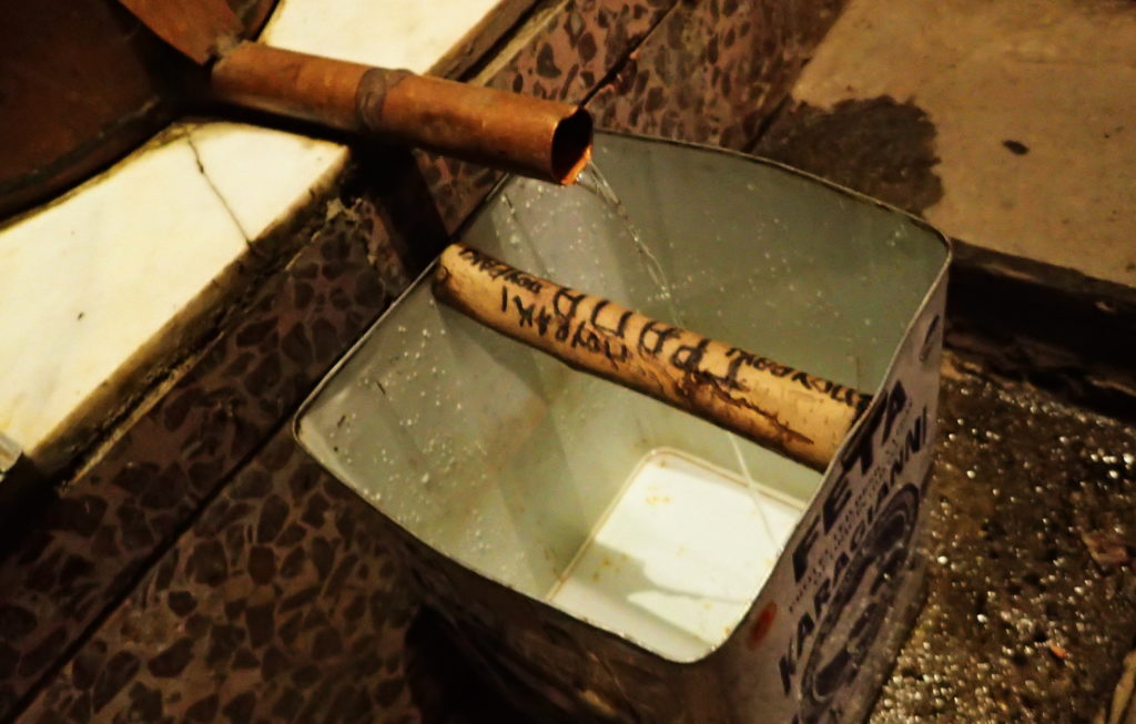 Tsipouro flows from the still into an old feta tin. The handle is marked "Grapa."